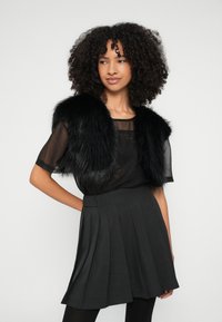 Colete de pelo sintético preto sobre uma blusa preta transparente, combinado com uma saia plissada cinza escuro e collants pretos. Texturas suaves, design elegante.