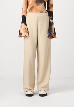 Pantalon beige à jambes larges en tissu lisse, avec une coupe droite et une taille minimaliste. Associé à un haut à motifs et des chaussures imposantes.