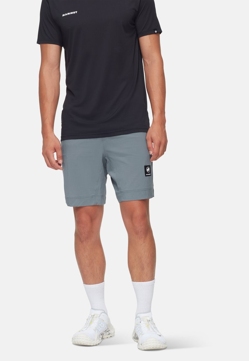 Herrklädsel bestående av en svart kortärmad skjorta och ljusgrå shorts med en svart logotyp, kombinerat med vita strumpor och sneakers.