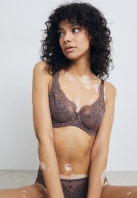 Soutien-gorge en dentelle marron avec des panneaux en maille transparente, motifs floraux et bords festonnés. Conception avec armatures et boutons décoratifs au centre.
