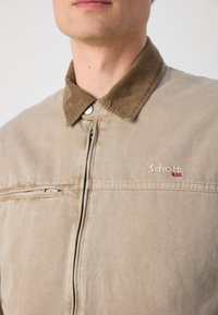 Chaqueta beige con cuello de pana marrón, cremallera frontal y bolsillo lateral con cremallera. Presenta el logo "Schott" en rojo en el área del pecho izquierdo.