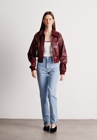 Μπορντό δερμάτινο bomber τζάκετ, λευκό φανελάκι, ανοιχτό μπλε τζιν παντελόνι, μαύρα παπούτσια. Το μοντέλο στέκεται απέναντι από ένα απλό φόντο.