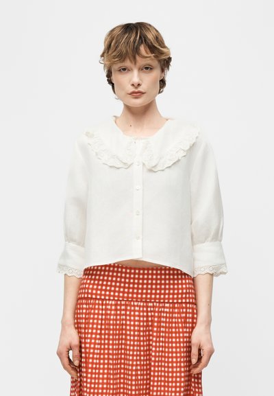 Mujer con una blusa blanca con un gran cuello y puños de encaje, combinada con una falda de cuadros rojos y blancos, posando frente a un fondo liso.