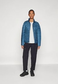 Mladić stoji, odjeven u plavu puf jaknu preko bijele košulje, crne hlače sa zip zatvaračima i crne tenisice na jednobojnoj pozadini.