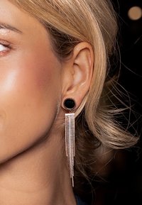 Boucles d'oreilles créoles noires avec une longue chaîne en rose gold. Le design présente une texture lisse et une forme géométrique de la chaîne.