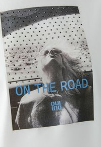 Witte t-shirt met een zwart-wit afdruk van een persoon met een zonnebril, versierd met zilveren studs. De tekst luidt "ON THE ROAD" in het blauw.