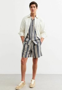 Camicetta a righe a maniche corte in blu e beige, abbinata a pantaloni corti a righe coordinati. Completata con una camicia overshirt color crema e sneakers beige.
