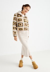 Chaqueta corta texturizada con patrones geométricos multicolores, cuello crema, combinada con una falda ajustada de color beige claro y zuecos de plataforma de ante marrón.