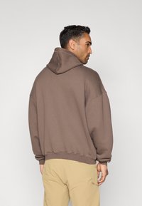 Sweatshirt castanha oversize feita de tecido texturizado, com capuz grande e punhos canelados. Vestida com calças bege, vista de costas.