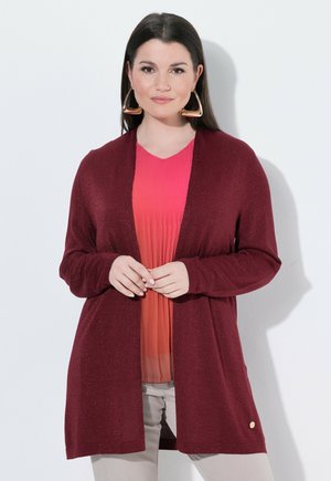 Vrouw met lang donker haar die grote driehoekige gouden oorbellen draagt, een rode cardigan over een roze geplooide top en een lichte broek.