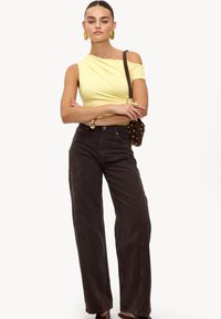 Haut asymétrique jaune à manches courtes, associé à un jean large taille haute marron foncé. Une bague simple et des boucles d'oreilles créoles sont des accents notables.