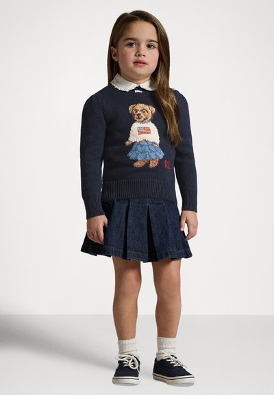 Temno modra pletena jopica z motivom medveda, belim ovratnikom in rdečimi poudarki, kombinirana s plisirano denim krilo in belimi nogavicami v modrih čevljih.
