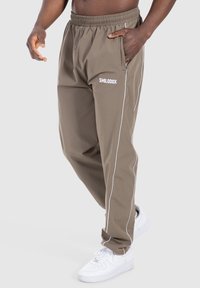 Pantaloni sportivi marroni con piping bianco sui lati, vita elastica e tasche con zip. Abbinali a delle sneakers bianche per un look casual.