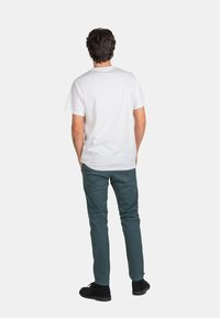 Camiseta blanca de manga corta y pantalones verdes. La camiseta tiene un cuello redondo; los pantalones tienen un ajuste slim. Zapatos negros con suela plana.
