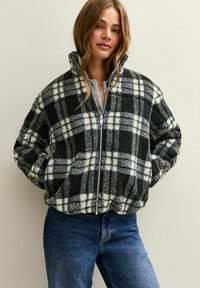Geruit jack met rits en een fuzzy zwart-witte textuur, met een hoge kraag en zijzakken. Draagt bij blauwe jeans.