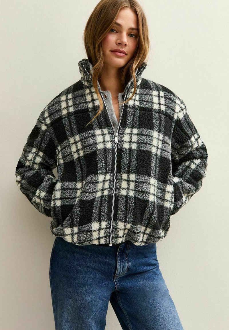 Geruit jack met rits en een fuzzy zwart-witte textuur, met een hoge kraag en zijzakken. Draagt bij blauwe jeans.