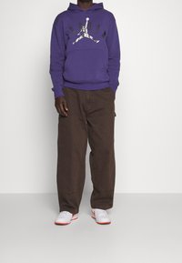 Sudadera morada con un logo de Jordan en negro y gris, combinada con pantalones sueltos marrones y zapatillas blancas con detalles en rojo.