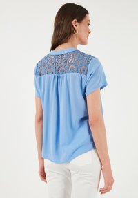 Blusa azzurra a maniche corte con pannello posteriore in crochet testurizzato e orlo arrotondato, realizzata in tessuto liscio. Pantaloni bianchi visibili qui sotto.