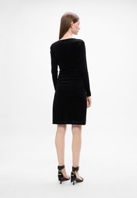 Čierne sametové bodycon šaty s dlhými rukávmi, priliehavým strihom, dĺžky po kolená a so zberaním na chrbte. Nosené s čiernymi vysokými sandálmi.