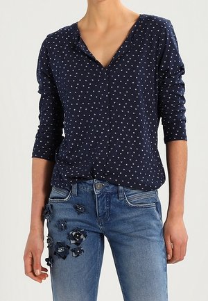Person iført en mørkeblå langærmet skjorte med små hvide prikker og blå jeans dekoreret med mørkeblå stofblomster på venstre lår.