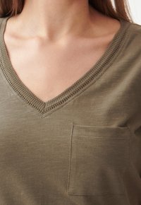 Olive green T-shirt z dekoltem w serek wykonany z miękkiego, strukturalnego materiału, z małą kieszonką na lewej piersi i ozdobnym szyciem wzdłuż dekoltu.