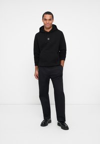 Hoodie noir avec poche avant et cordons, arborant un petit logo blanc. Associé à un pantalon noir et des chaussures noires.