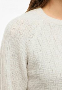 Gaiši krēmkrāsas džemperis ar strukturētu rakstu, apaļu kakla izgriezumu un raglāna piedurknēm. Materiāls izskatās mīksts un austs ar niansētu apdari.