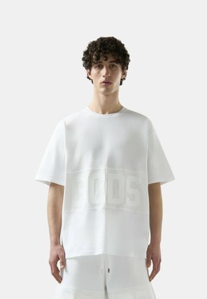 Giovane con capelli ricci che indossa una maglietta bianca oversize con grandi lettere "GCDS" in rilievo e pantaloncini bianchi abbinati.