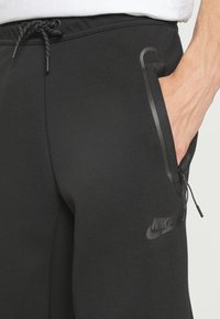 Černé Nike tepláky s elastickým pasem, stahovací šňůrkou, zipovými bočními kapsami a tónovaným logem. Hladká tkanina s přiléhavým střihem.