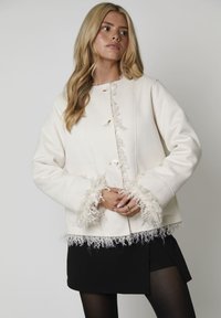 Kurtka w kolorze ecru z miękkim, teksturowanym frędzlem wzdłuż mankietów i dołu. Posiada okrągłe guziki, prosty krój oraz minimalistyczny design.