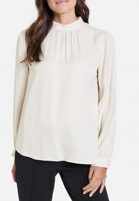 Blusa lunga color beige chiaro con collo alto e dettagli arricciati. Tessuto liscio, design fluido e vestibilità leggermente ampia.