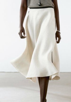 Femme portant une jupe midi blanche fluide avec une fente latérale, associée à un haut gris et un pendentif noir sur un collier.