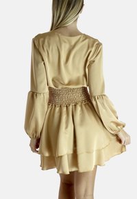 Abito A-line in satin di un morbido beige con maniche lunghe a sbuffo, vita arricciata e orlo a strati, che offre una texture liscia e una silhouette fluida.