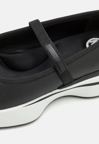 Schwarzer synthetischer Schuh mit elastischem Riemen, strukturiertem Obermaterial und gepolsterter Innensohle. Weiße Sohle mit schwarzem Akzent. Logo prägegedrückt auf dem Riemen.