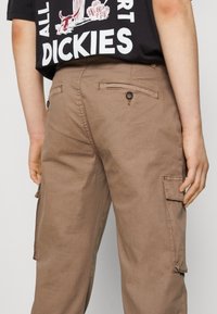 Persona vistiendo pantalones cargo marrones con bolsillos traseros abotonados y una camiseta gráfica negra con el texto "DICKIES" en blanco.