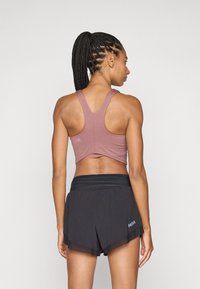 Sporte atvars mauve krāsā ar sacensību aizmuguri, kombinēts ar melniem šortiem, kas izceļas ar slāņu izskatu un logo detaļu kreisajā pusē.