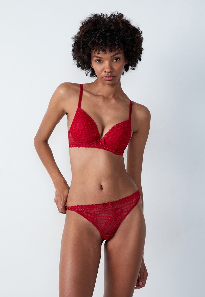 Soutien-gorge en dentelle rouge avec un design triangulaire, des bords festonnés et un petit nœud au centre. String en dentelle assorti avec un motif similaire.
