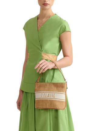 Donna con indosso un vestito verde che tiene una borsa intrecciata color cammello con una striscia bianca e la scritta "1 CLASSE", vista dal collo ai fianchi.