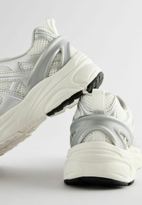 Witte sportschoenen met gaaspanelen, textuuraccenten, een grijze hielbeugel en een zwarte rubberen zool. Kenmerkt een trekband aan de hiel.