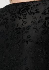 Μαύρο ύφασμα με λουλουδάτο σχέδιο και ανάγλυφη υφή βελούδου. Το μοτίβο περιλαμβάνει τριαντάφυλλα και φύλλα, δημιουργώντας ένα διακριτικό, πολυεπίπεδο εφέ.