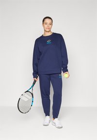 Tuta sportivo blu navy realizzato in tessuto morbido, con logo sulla felpa e sui pantaloni. Il modello tiene in mano una racchetta da tennis e una pallina.