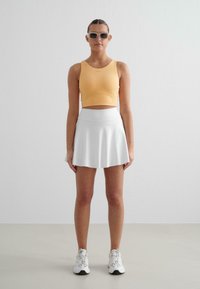 Koorangist lühike tank top, valgest voolav seelik ja valged spordijalatsid. Mudel kannab halli ristkülikukujulist prille sujuva tekstuuriga taustal.