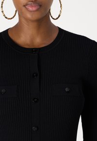 Fekete bordázott kötött kardigán kerek nyakkivágással, elülső gombokkal és két zsebbel a mellrészen, amelyeken gombos zárás található.