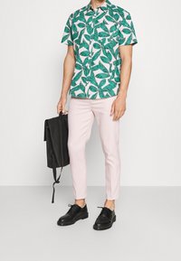 Uomo che indossa una camicia a maniche corte con motivo a foglie verdi, pantaloni rosa chiaro, scarpe nere, che tiene uno zaino nero per il manico, in piedi su uno sfondo neutro.