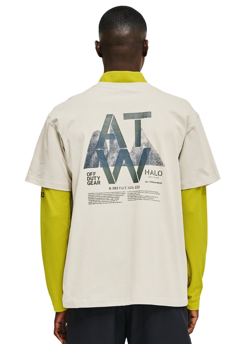 Beżowa koszulka z krótkim rękawem z dużym nadrukiem z zielonym i niebieskim tekstem z tyłu, a pod spodem żółta siateczkowa koszulka z długim rękawem.