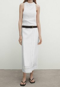 Biała top bez rękawów z wysokim dekoltem, zestawiona z białą spódnicą midi z wycięciami, podkreślona czarnym paskiem i klapkami.