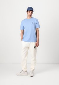 T-shirt en coton bleu clair avec logo imprimé ; sweatpants crème avec texte noir ; baskets grises ; casquette bleue. Le modèle se tient devant un fond uni.