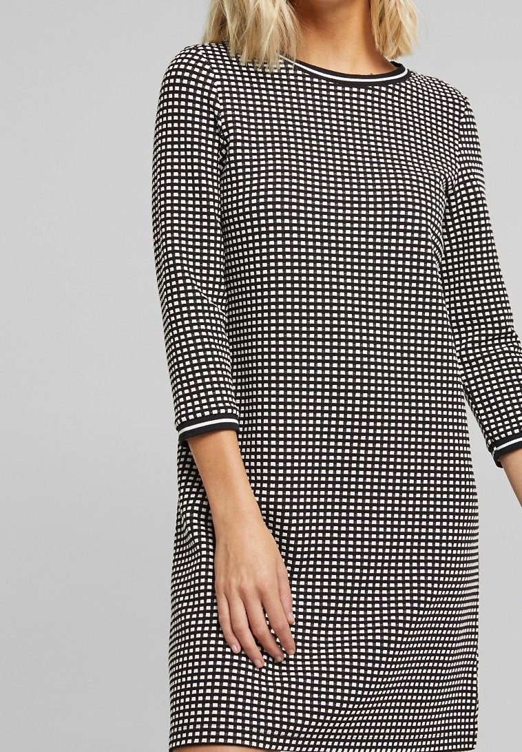 Femme portant une robe à carreaux noir et blanc avec des manches trois-quarts et un col rond sur un fond gris uni.