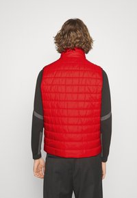 Rote gepolsterte Weste mit hohem Kragen, gesteppter Textur und schlankem Design. Getragen über einem langen grauen Shirt und dunklen Hosen.