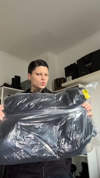 Vestuário preto embalado em plástico transparente com uma etiqueta amarela. A textura visível sugere material macio. O fundo inclui calçado preto e bolsas.